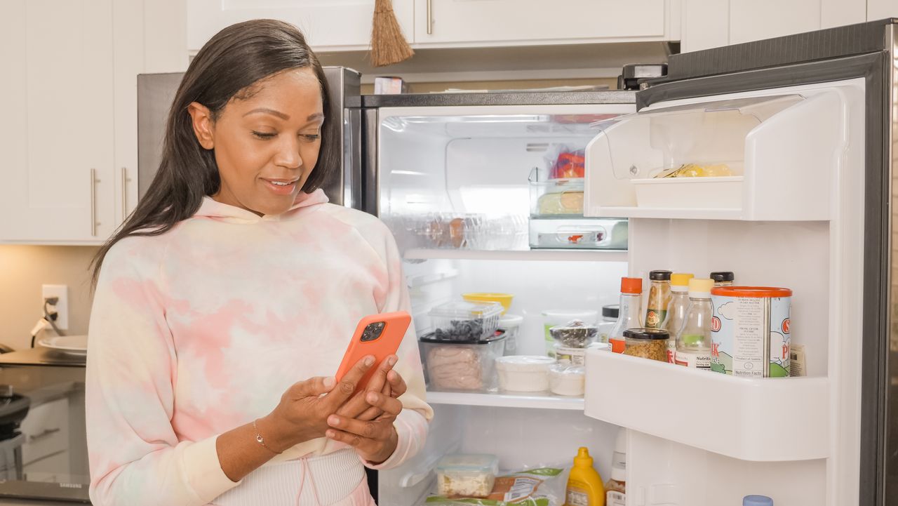 Woman using smartphone for online shopping while standing in the kitchen with an open refrigerator.